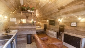 A cozy cabin interior with a wooden interior, a fireplace, a sofa, and a kitchen area with a kettle and utensils.