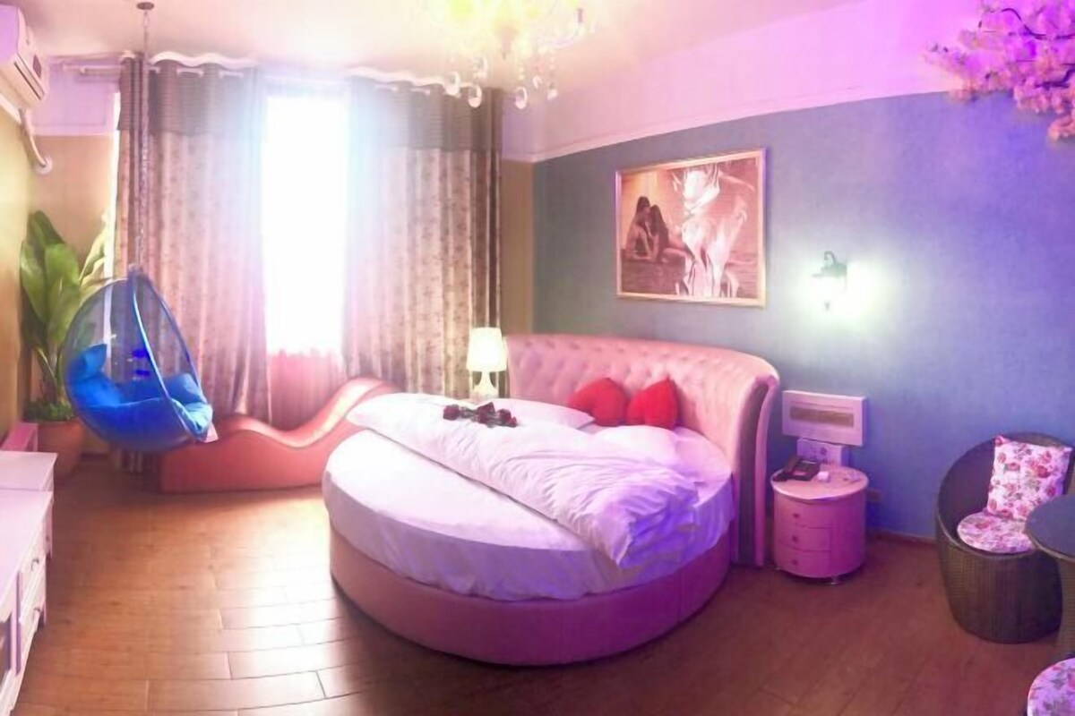 Een slaapkamer met een rond bed, een hangstoel, een nachtkastje met lamp en een aan de muur bevestigde televisie.