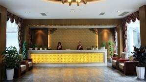 Uma área de recepção com balcão, dois recepcionistas, um candelabro e plantas em vasos.