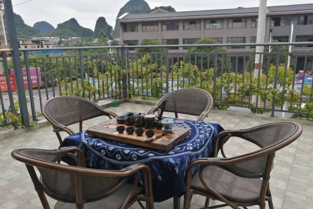 A balcony with a table set for tea, surrounded by wicker chairs, overlooking a garden and buildings.