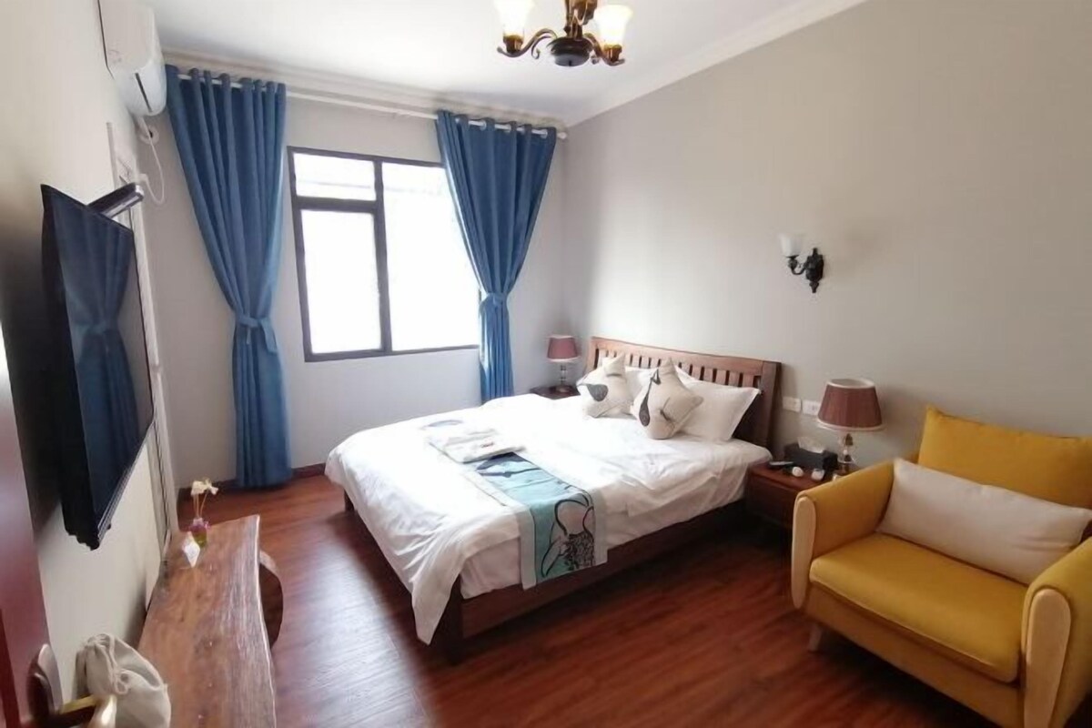 A hotel room with a bed, a yellow armchair, a bedside table with lamps, a window with curtains, and a flat-screen TV mounted on the wall.