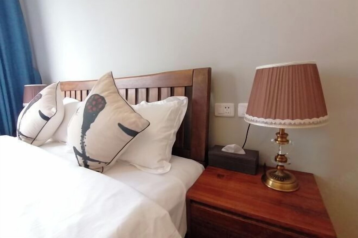 A neatly made bed with white bedding and patterned pillows, a wooden nightstand with a lamp, and a power outlet on the wall.