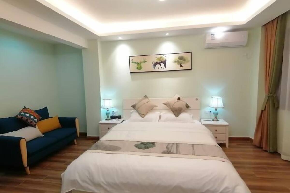 A bedroom with a large bed, two bedside tables with lamps, a blue sofa with colorful cushions, and a wall-mounted air conditioner.