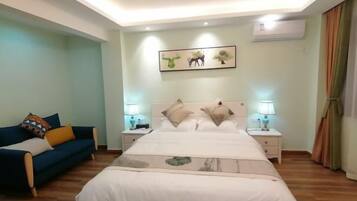 A bedroom with a large bed, two bedside tables with lamps, a blue sofa with colorful cushions, and a wall-mounted air conditioner.