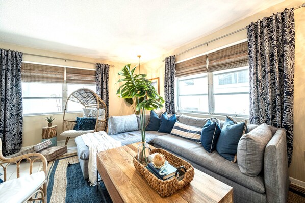 A cozy living room with a sofa, a coffee table, and a chair. There is a plant on the coffee table and a window with curtains.