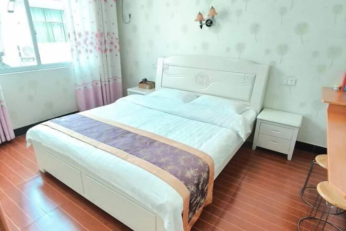 A bedroom with a bed, bedside tables, a nightstand, a window with curtains, and a wall-mounted lamp.