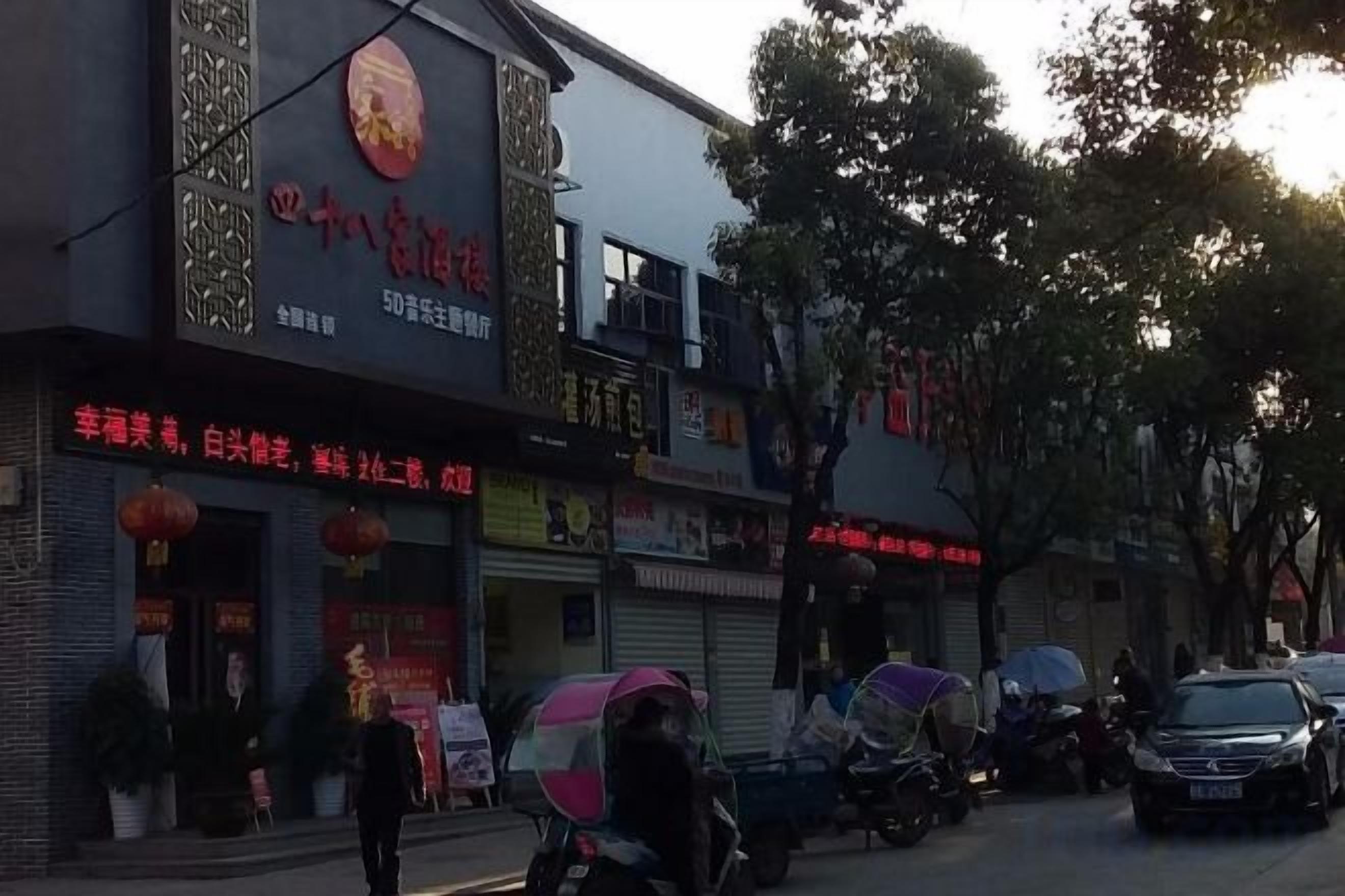 A street view with a building featuring a large sign with Chinese characters, a storefront with a red awning, and parked vehicles.