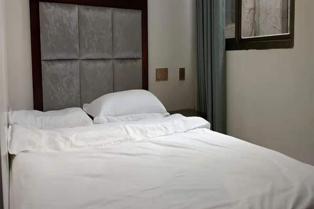 A neatly made bed with white linens in a room with a padded headboard and a window with curtains.