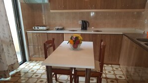 Une cuisine avec un sol carrelé, une table blanche avec des chaises, un panier de fruits et une bouilloire posée sur le comptoir.