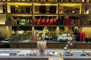 Un espace bar bien aménagé, avec des étagères remplies de différentes bouteilles et de verres, un comptoir où sont exposés des articles à vendre, et une vue dégagée sur l’espace bar à travers la vitre.