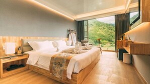 Ein modernes Hotelzimmer mit einem großen Bett, Holznachttischen, einem Beistelltisch, Blick auf Grün durch das Fenster und einer eingebauten Ablagefläche.