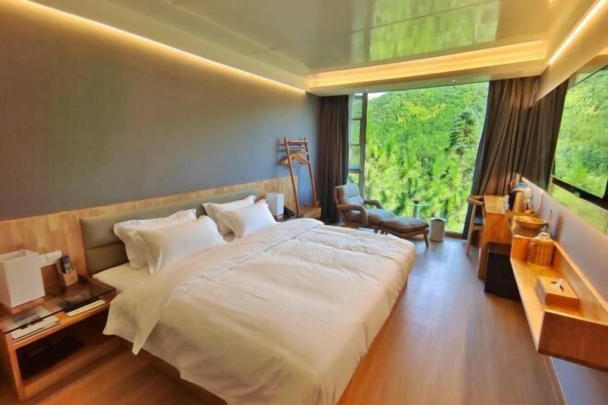Ein modernes Hotelzimmer mit einem großen Bett, einem Schreibtisch, einem Stuhl und Blick auf Grün hinter dem Fenster.