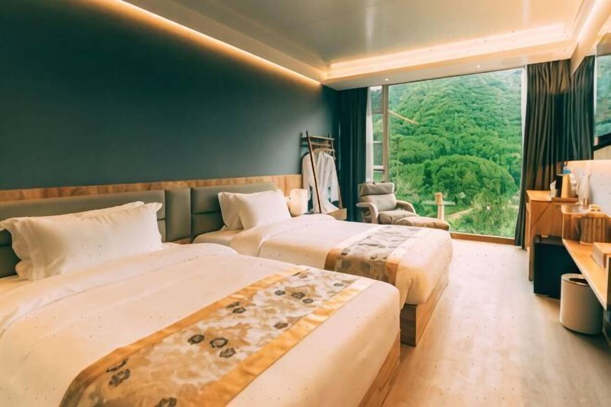 Ein Hotelzimmer mit zwei Betten, einem Schreibtisch, einem Sessel und einem großen Fenster mit Blick auf Grünflächen.