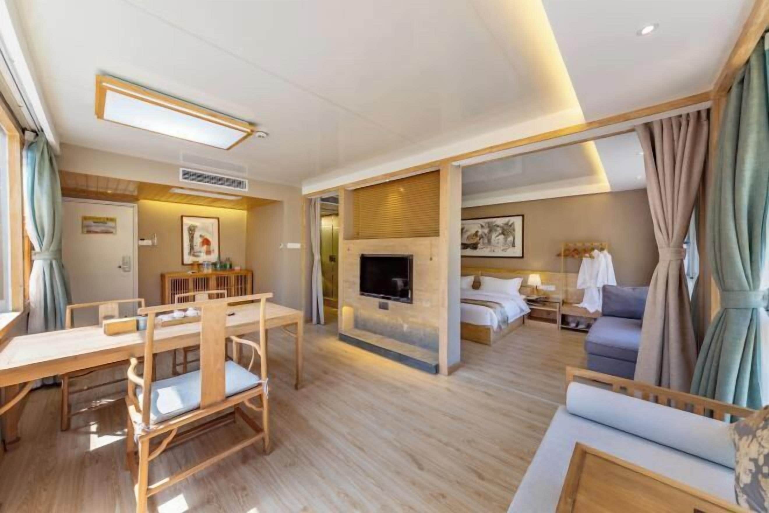 Habitación de hotel con una mesa de comedor de madera, un sofá, un televisor montado en la pared y una cama con una mesita de noche.
