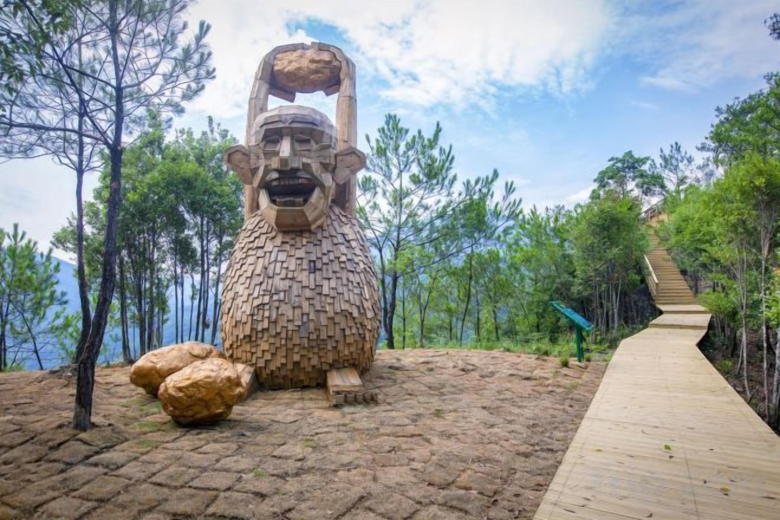Une sculpture représentant une figure assise, avec une grande tête et un corps plus petit, entourée d’arbres et d’un sentier en bois.