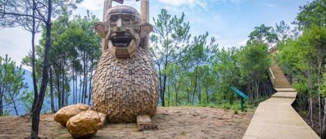 Une sculpture représentant une figure assise, avec une grande tête et un corps plus petit, entourée d’arbres et d’un sentier en bois.