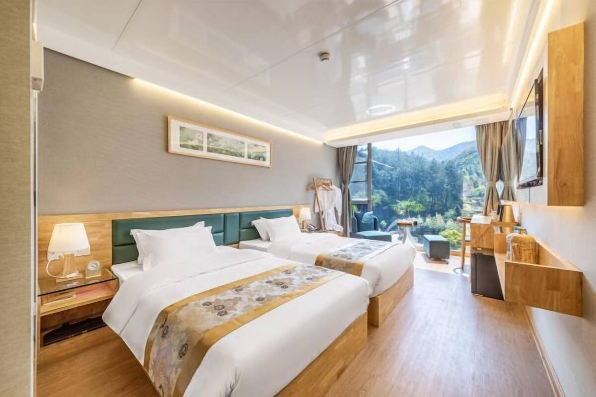 Ein Hotelzimmer mit zwei Betten, einem großen Fenster mit Blick auf Grünflächen, einem Flachbildfernseher und einem Nachttisch aus Holz mit einer Lampe.
