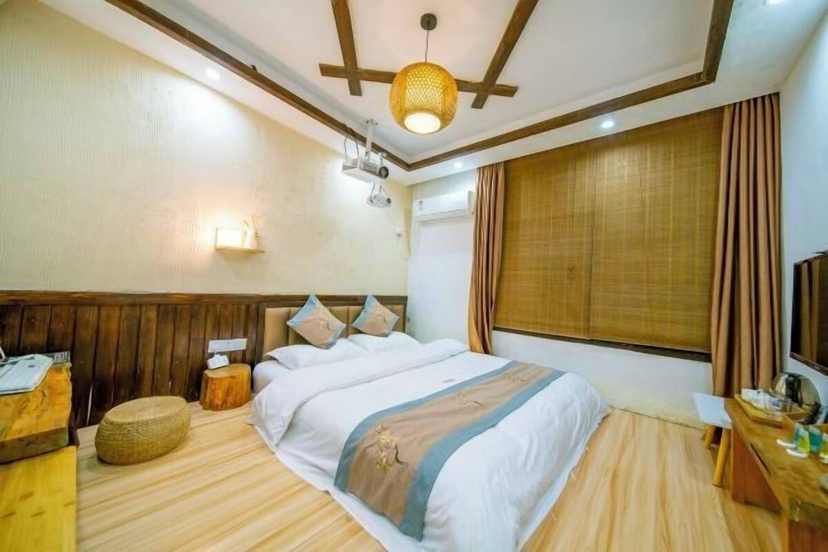 Habitación de hotel con cama, piso de madera, escritorio, lámpara, luz en la pared y ventana con cortinas.