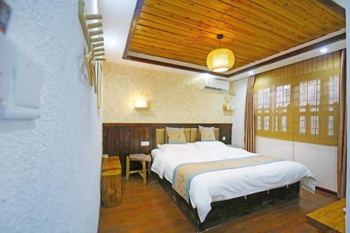 Un dormitorio con techo de madera, cama con ropa blanca, un taburete pequeño y una ventana con cortinas.