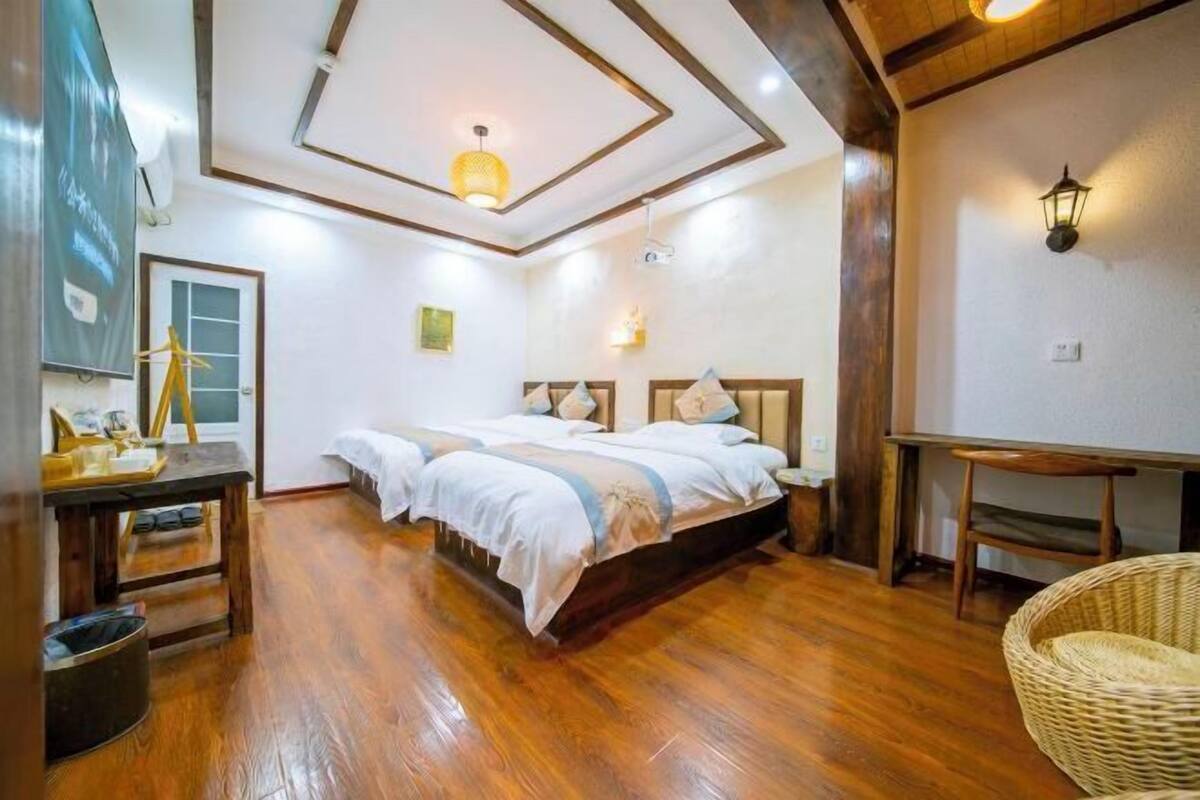 Una habitación con piso de madera, una cama con ropa blanca, una mesita, una silla de mimbre y una lámpara fijada en la pared.
