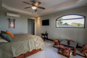Un dormitorio con cama, ventilador de techo, televisor, ventana y una pequeña mesa con sillas.