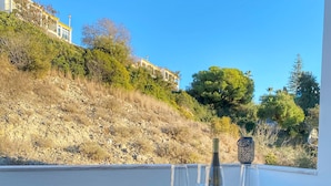 A glass table with chairs and plates, a bottle, and glasses on a balcony with a view of a hill and trees.