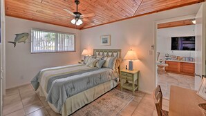 A bedroom with a wooden ceiling, a bed with a headboard, two bedside lamps, a TV, and a small dining area with a table and chairs. - Everglades Getaway w/ Deck & Water Views! (Everglades City)