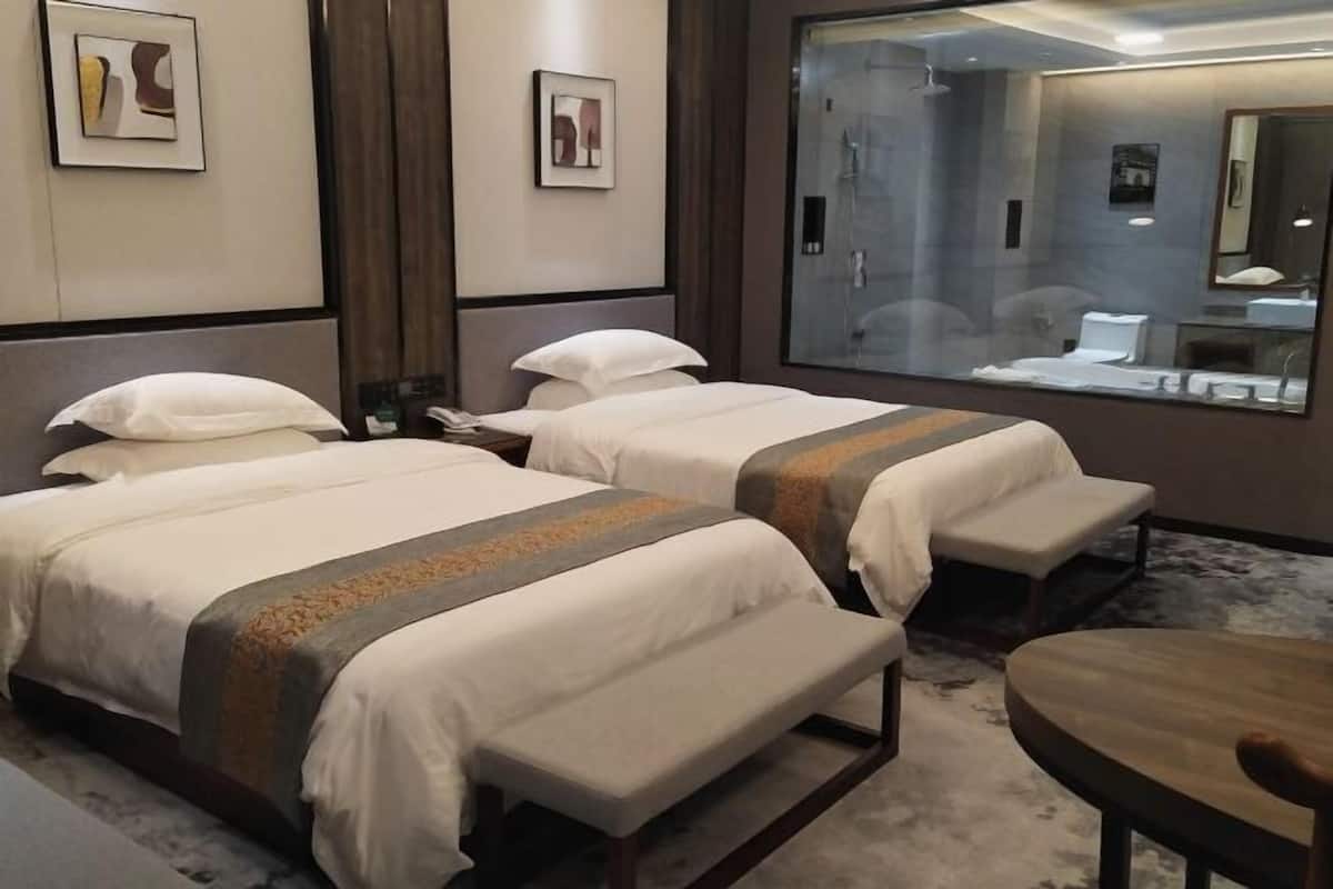 Habitación de hotel con dos camas, un banco, una mesa de madera y un espejo grande que refleja el baño.