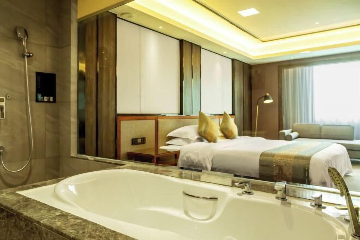 A modern hotel room with a large bathtub, a bed with pillows, a bedside table, and a large mirror reflecting the room.