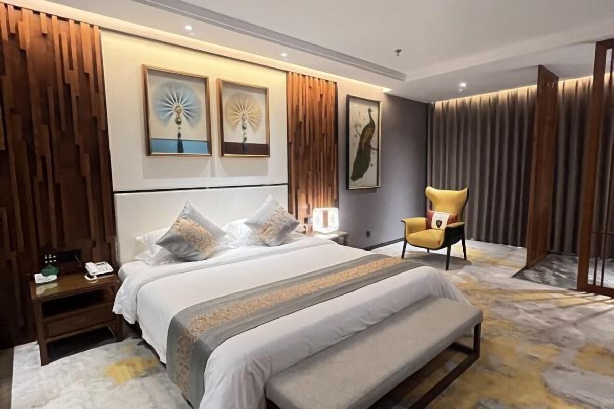A modern hotel room with a large bed, a bedside table with a lamp, a chair, and artwork on the walls.