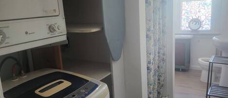A laundry area with a washing machine and dryer, a small window with a floral curtain, and a view of a bathroom with a toilet and sink.