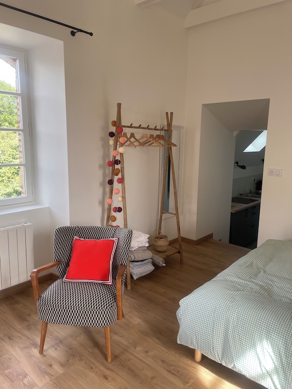 A room with a wooden floor, a patterned armchair with a red cushion, a bed with a patterned bedspread, a wooden clothes rack with hanging clothes, and a window with a view of greenery.