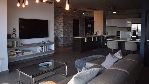 A modern living room with a sofa, coffee table, and TV, a kitchen with bar seating, and a stone accent wall. - Modern Condo at Atlantis (San Juan)