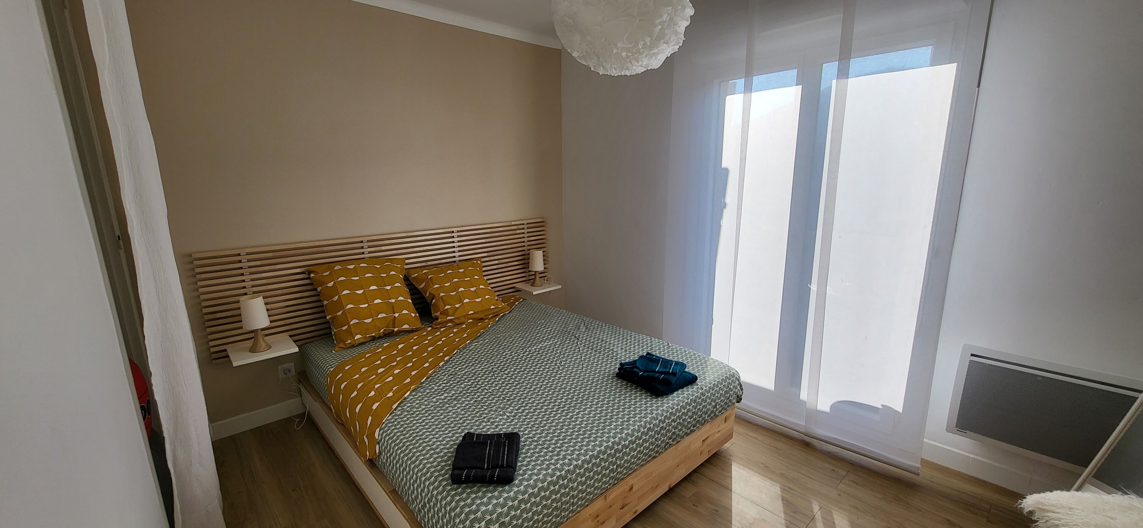 Une chambre à coucher avec un lit, une tête de lit en bois, des lampes de chevet, une fenêtre avec des stores et un radiateur.