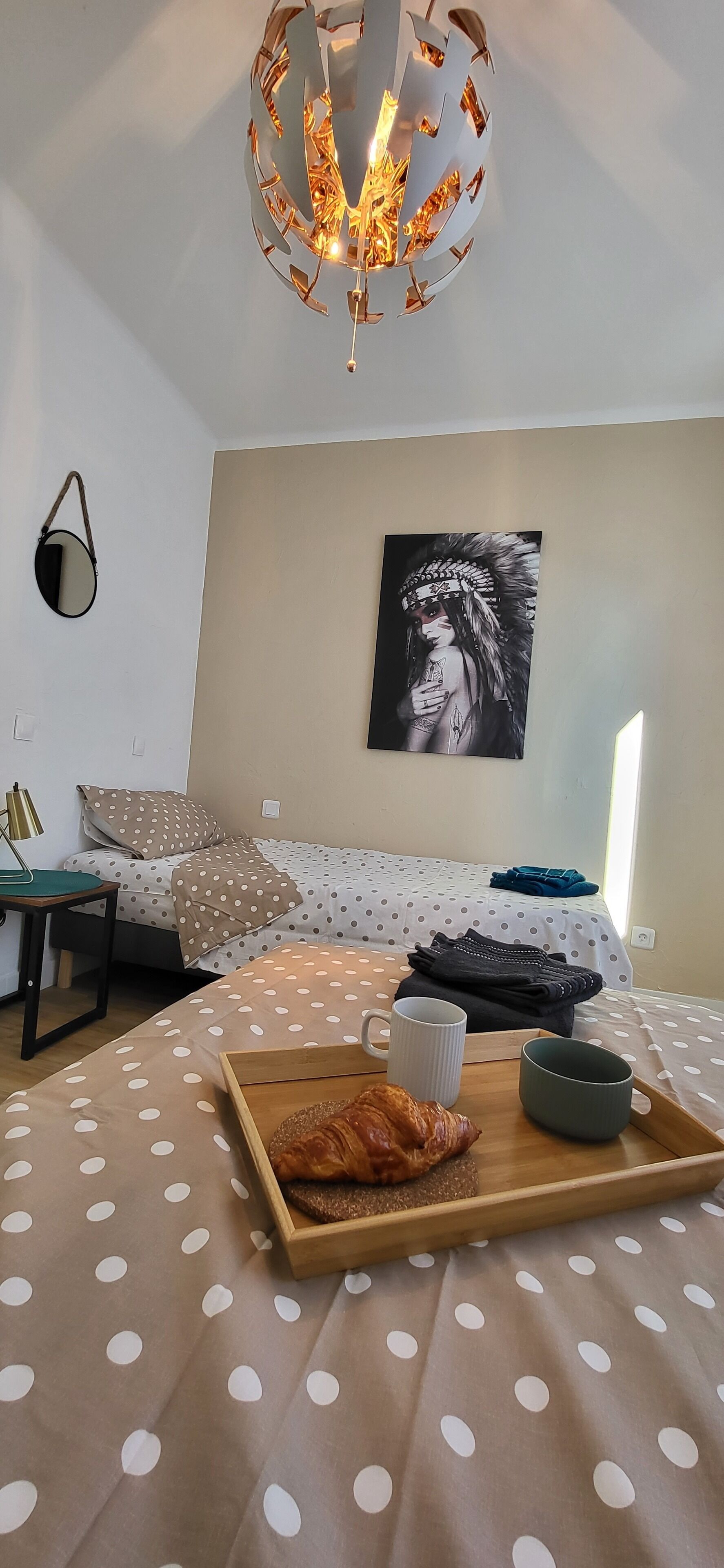 A bedroom with a polka dot bedspread, a wooden tray with croissants, a cup, and a painting on the wall.