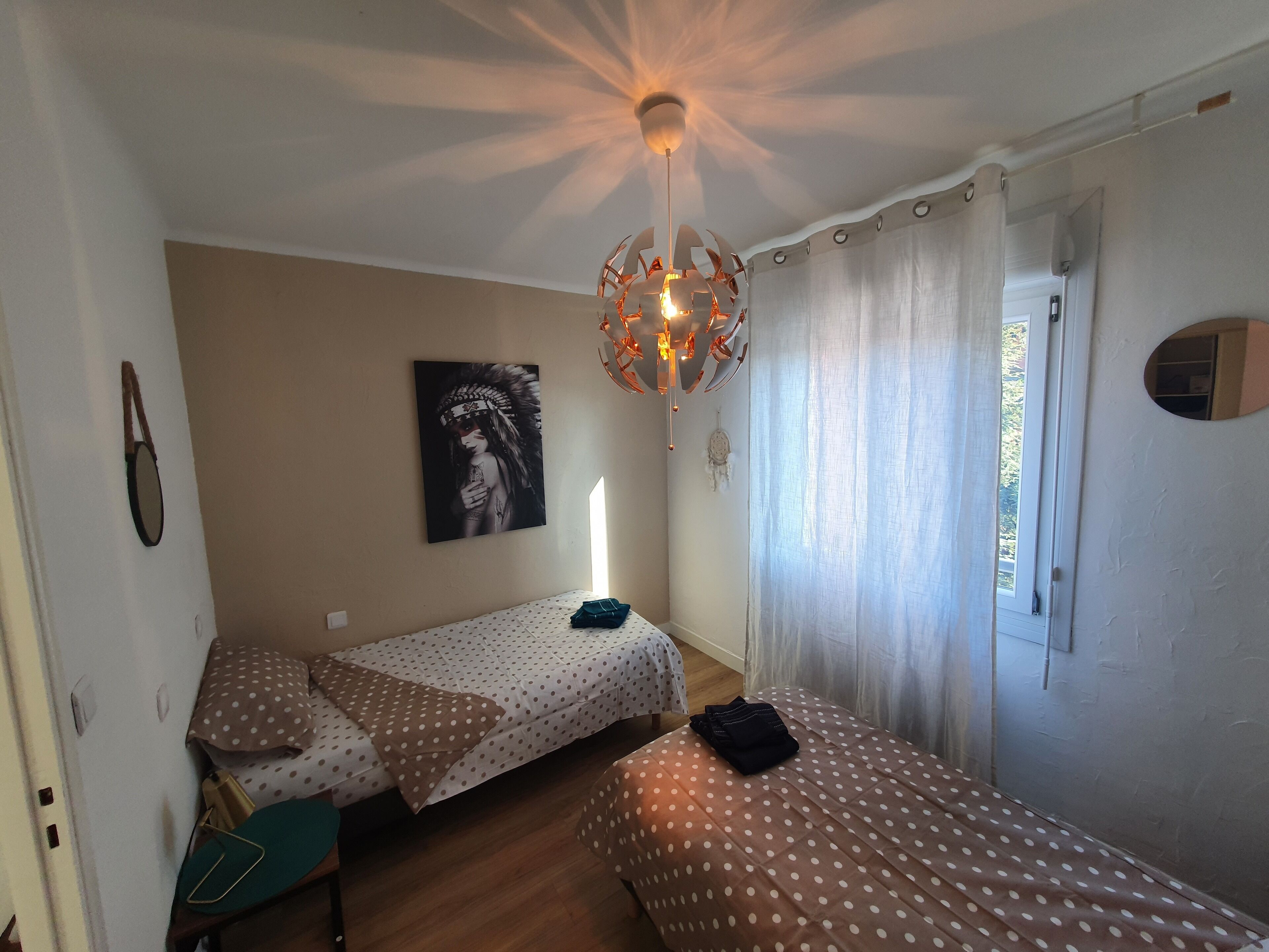 Una habitación con dos camas, una lámpara en el techo, un cuadro colgado en la pared y una ventana con cortinas.