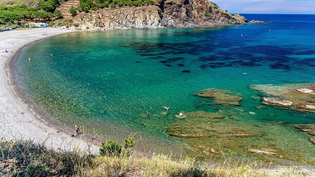 Ein Küstenstrand mit klarem türkisblauem Wasser, Felsformationen und einer hügeligen Landschaft im Hintergrund.