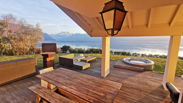 Een houten terras met een tafel en banken, uitkijkend over een meer met bergen op de achtergrond.