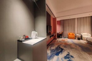 Una habitación de hotel moderna con pared gris, lavamanos blanco, alfombra estampada, sillón naranja y sofá blanco.