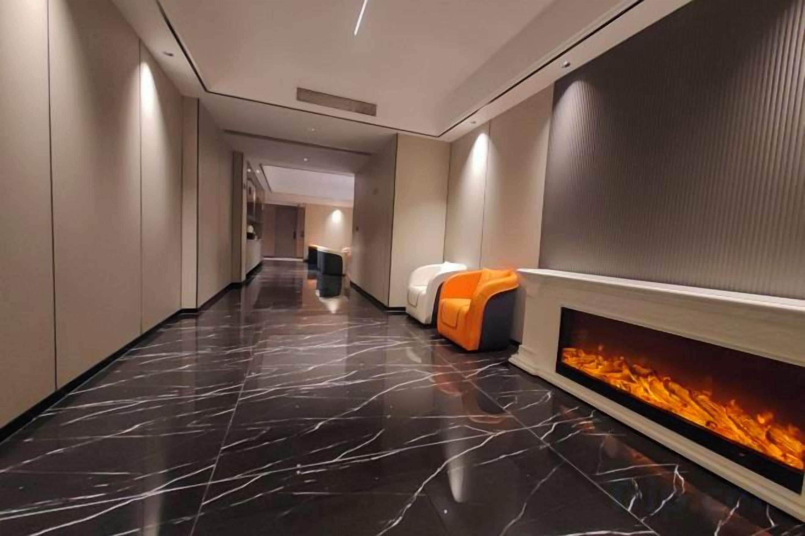 Un pasillo de hotel con piso de mármol negro, una zona de estar con mobiliario naranja y blanco, y una chimenea empotrada en la pared.