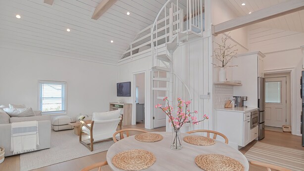 A spacious living area with a high ceiling, a dining table with placemats, a staircase, and a kitchen area. - The Beach Nest-Lux Cottage Steps to the Beach (Clinton)