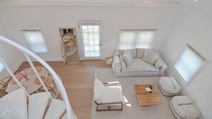 A spacious living area with a white sofa, a coffee table, and a dining table with chairs. There are two windows with blinds, a door, and a staircase leading to an upper level. - The Beach Nest-Lux Cottage Steps to the Beach (Clinton)