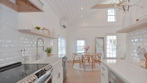 A modern kitchen with white cabinetry, a stainless steel oven, and a dining area with a wooden table and chairs. - The Beach Nest-Lux Cottage Steps to the Beach (Clinton)