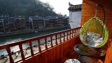A wooden balcony with a hanging chair, a round table, and a view of traditional buildings across a river.