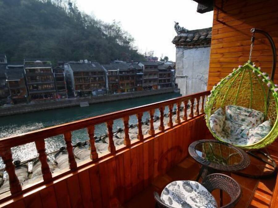A wooden balcony with a hanging chair, a round table, and a view of traditional buildings across a river.