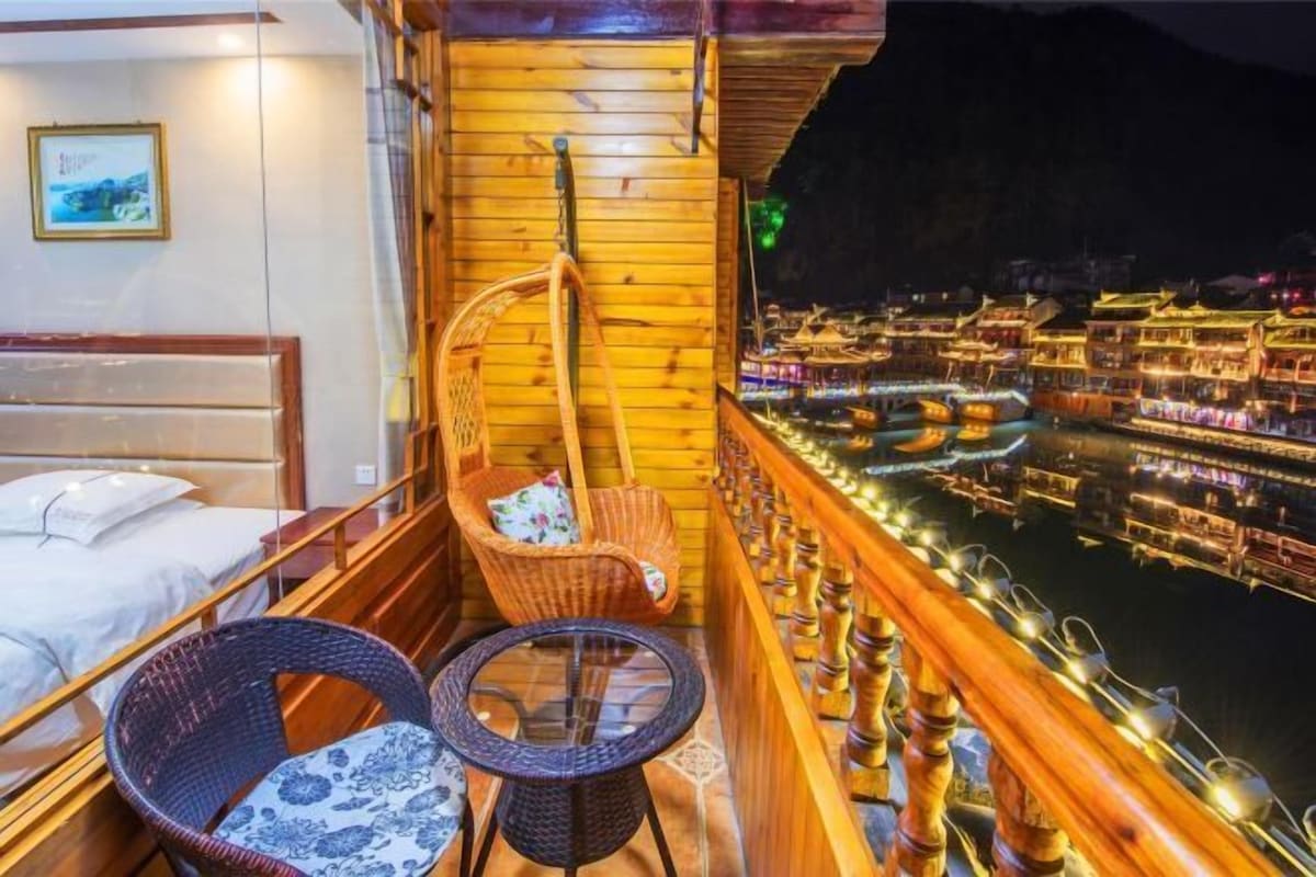 A balcony with wicker chairs and a small table, overlooking a lit-up riverside with traditional buildings.