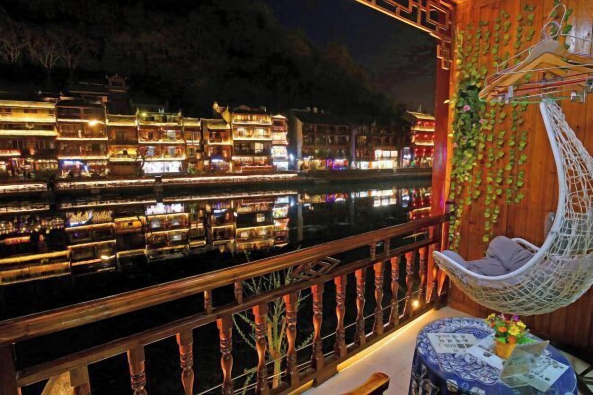 A balcony with a hanging chair, a table with a vase, and a view of illuminated buildings across the water.