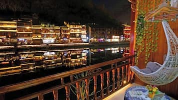 Un balcón con una silla colgante, una mesa con un florero y vistas a edificios iluminados a través del agua.