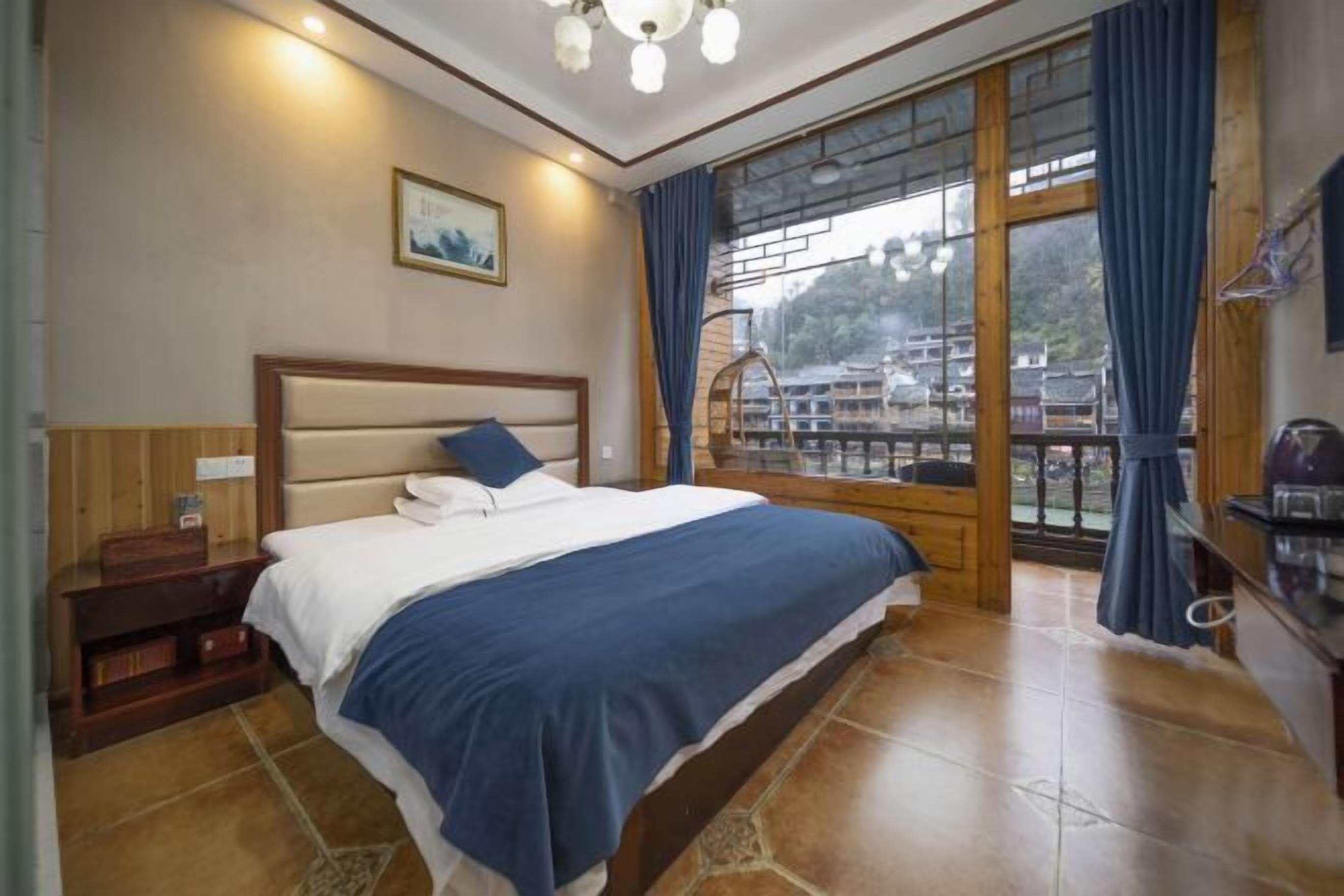 A bedroom with a bed, wooden headboard, a nightstand, a window with curtains, and a view of buildings outside.
