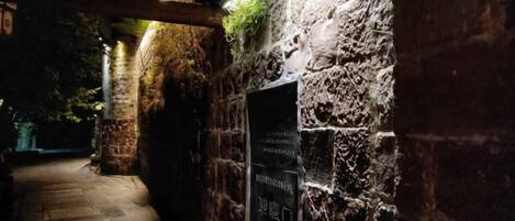 Un muro de piedra con una placa, una viga de madera en el techo y un sendero que se adentra en la distancia.